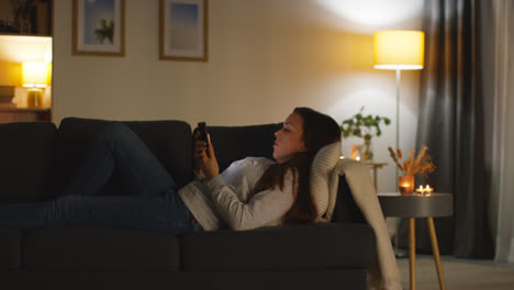 Woman-Lying-On-Sofa-At-Home-At-Streaming-Or-Watching-Movie-Or-Show-Or-Scrolling-Internet-On-Mobile-Phone